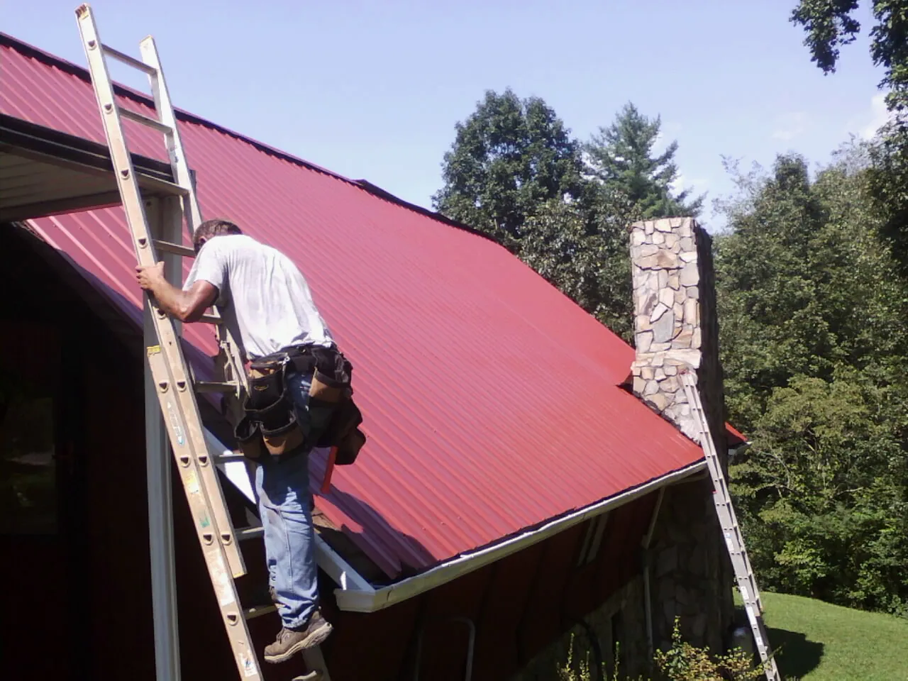 Roofer on a ladder beside a red metal roof and stone chimney — Soffit & Fascia Repair by Roofing Brothers in Hendrix